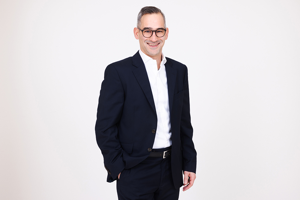 Portrait von Adrian Martinez mit weissem T-Shirt, Brille und Bart, Kurze schwarz-graue Haare